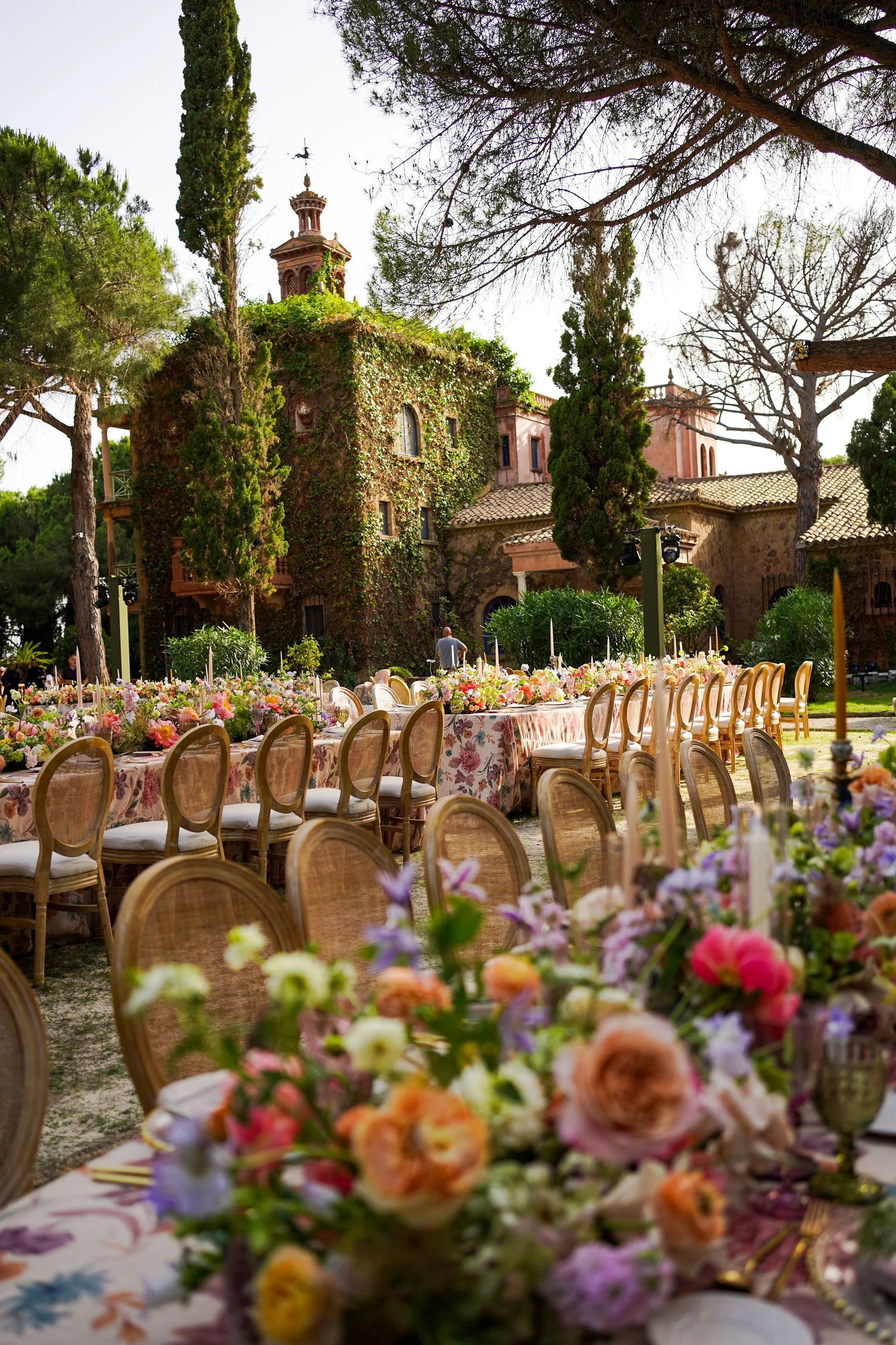 An outdoor event setup with long tables decorated with floral arrangements, surrounded by chairs, in front of an old ivy-covered building with a tower and tall trees.