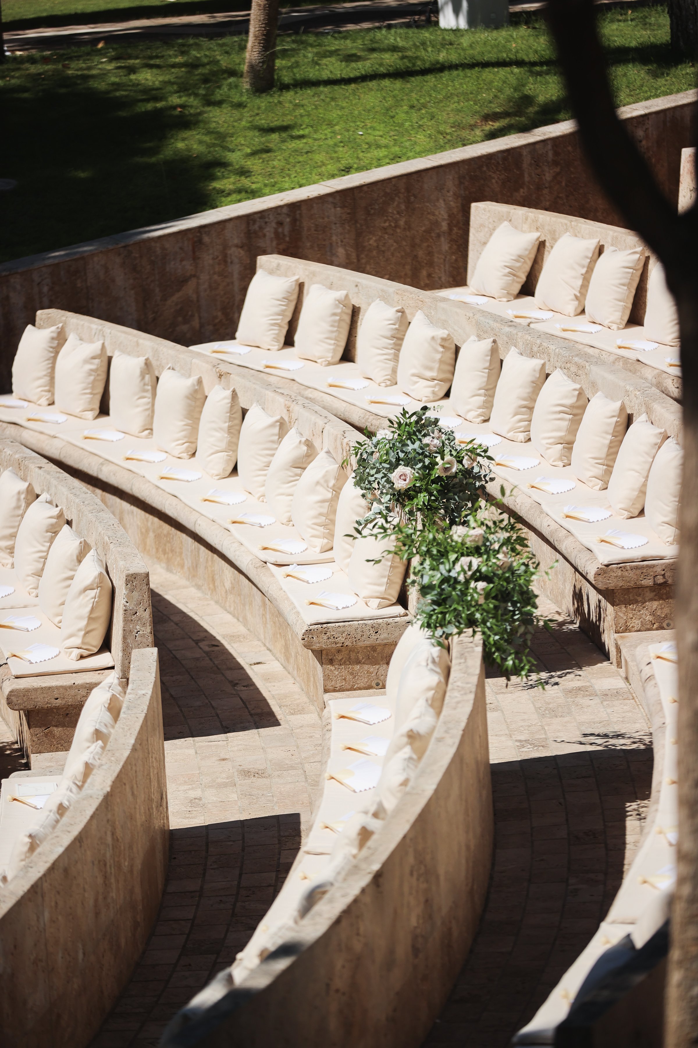 Empty tiered outdoor seating area with cushioned benches and a small floral arrangement in the foreground, with a grassy area in the background.
