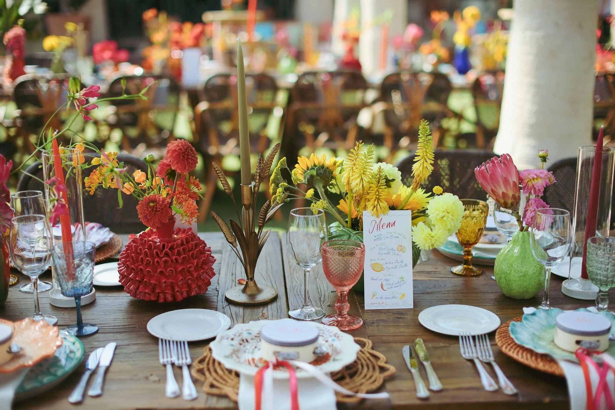 A colorful table setting with various decorated dishes, drinking glasses, and a menu card, featuring vibrant flowers as centerpieces, set outdoors for a first night wedding party at Anantara Villa Padierna Resort in Marbella