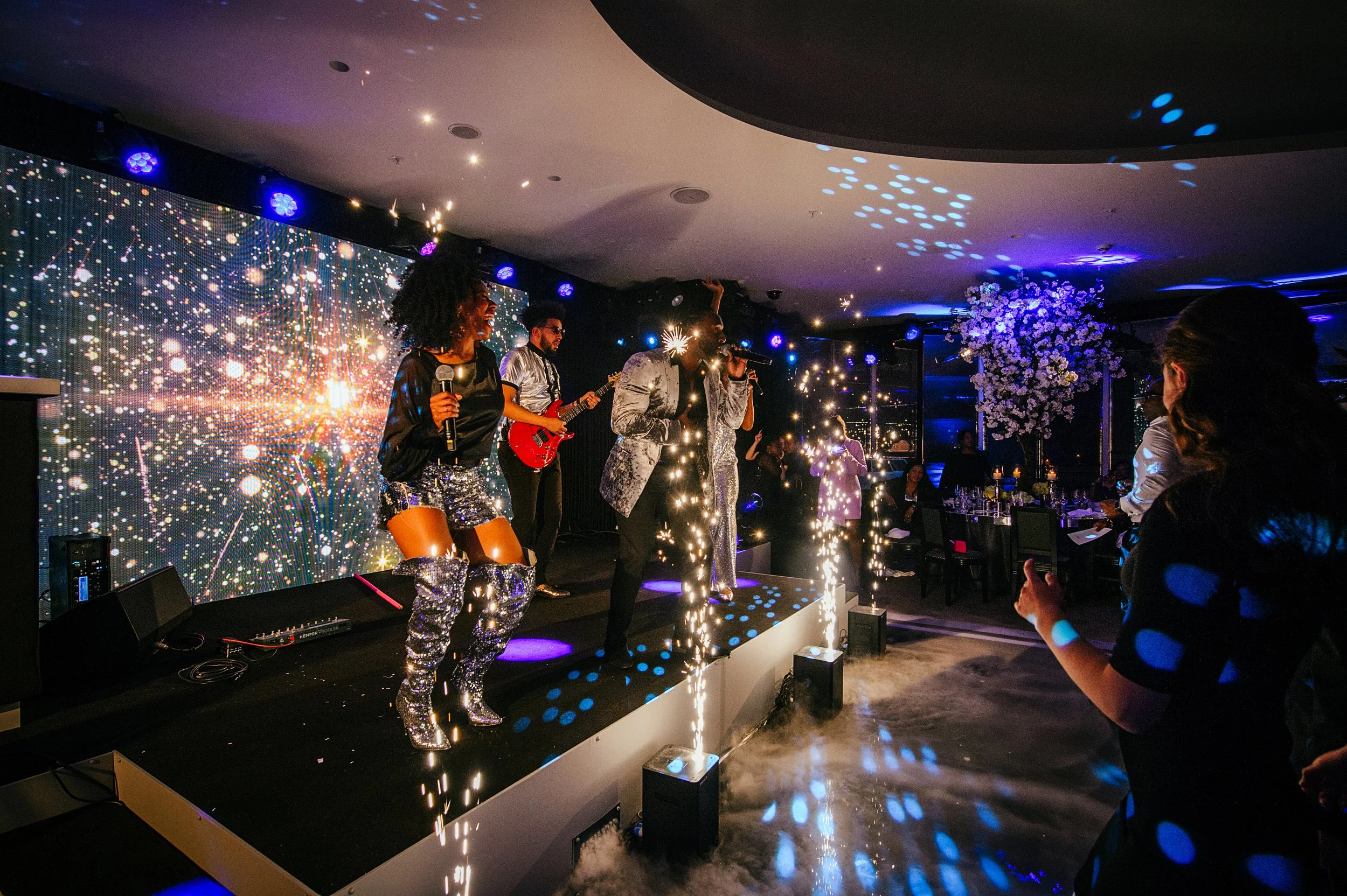 A lively stage performance with multiple musicians, including a woman holding a microphone, a guitarist, and a singer, with sparkling lights, pyrotechnics, and a digital backdrop of fireworks, in a darkened venue with audience members taking photos.