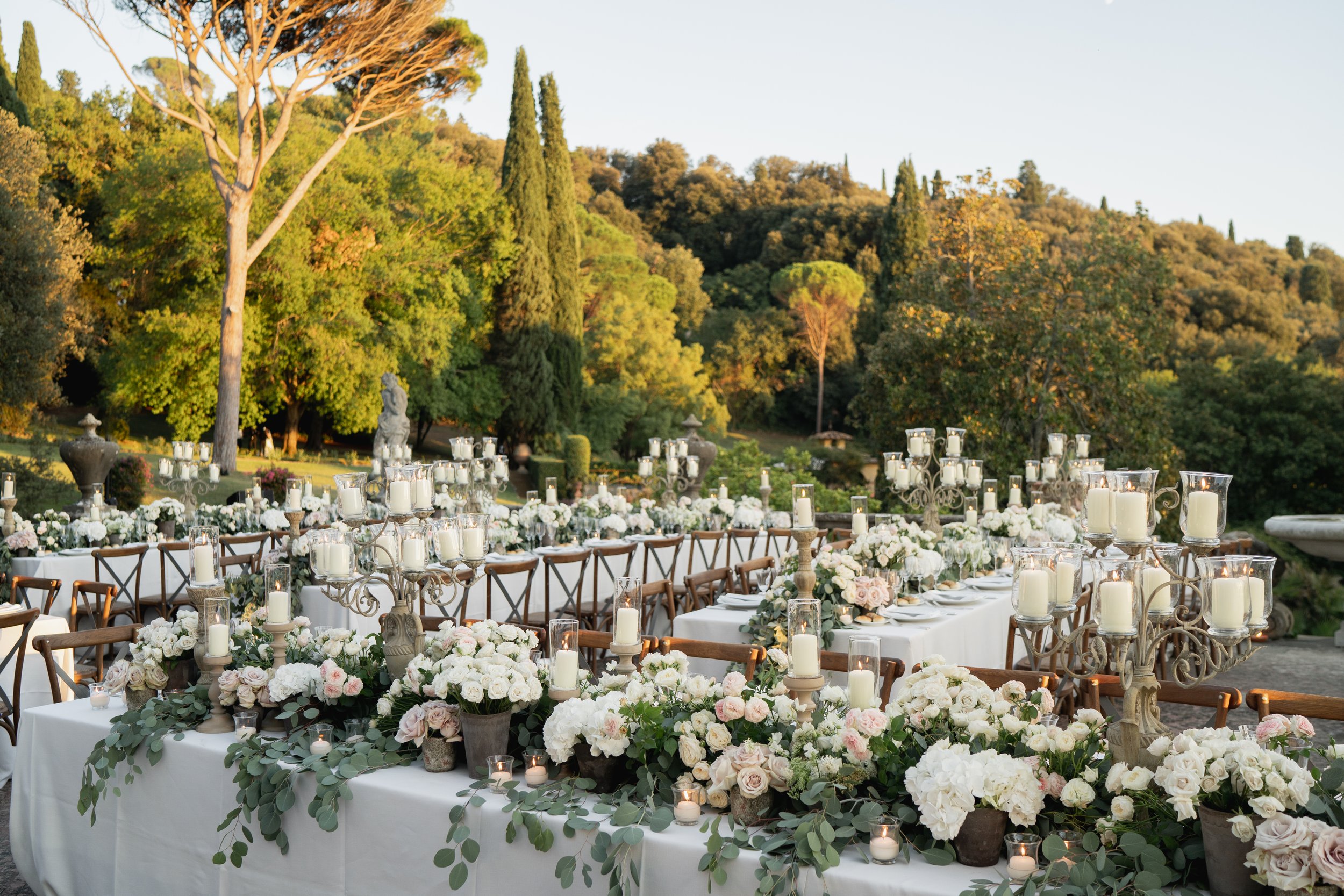 Outdoor event setup with decorated tables, numerous candles, and floral arrangements in a lush garden with tall trees and hills in the background at Villa Palmieri in Florence