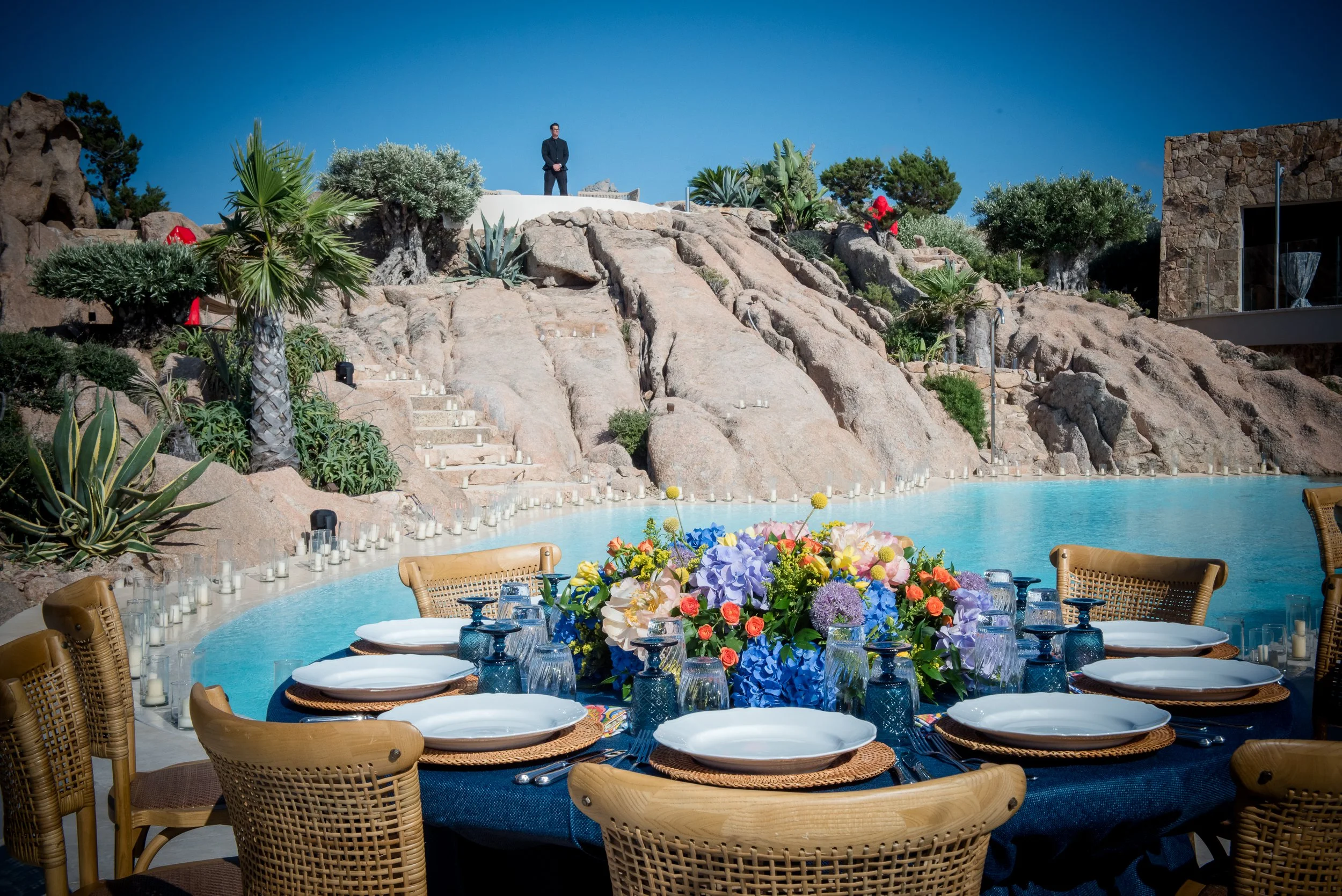 A lavish outdoor dining setup with a long table adorned with a vibrant floral centerpiece, set with plates, glasses, and silverware, near a swimming pool surrounded by desert landscaping with rocks, cacti, and succulents, under a clear blue sky, with