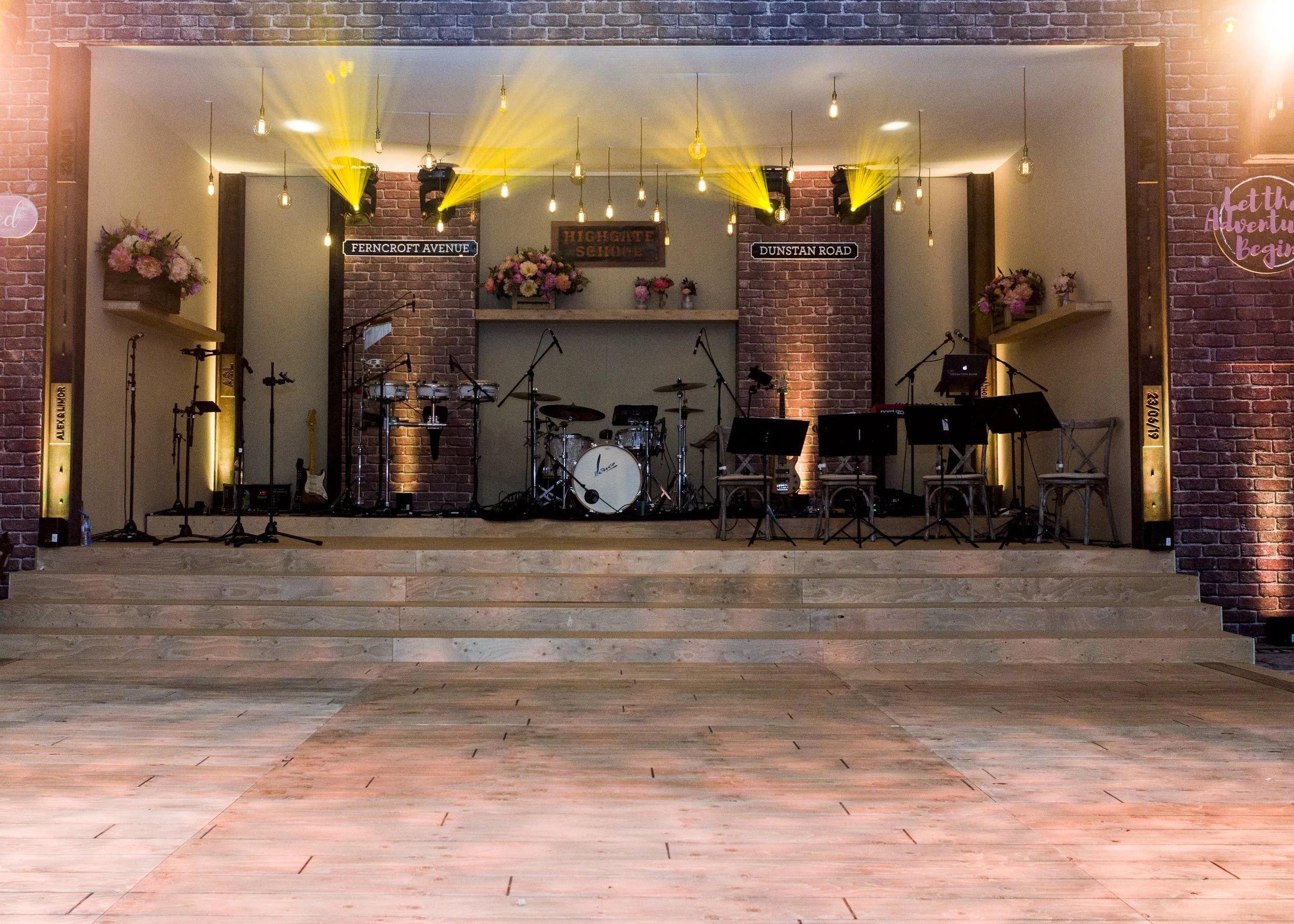 An indoor stage setup with musical instruments including drums and microphones, decorated with pink and white flowers, and illuminated by yellow stage lights, with signs reading 'Ferncroft Avenue' and 'Dunstan Road' on brick walls.