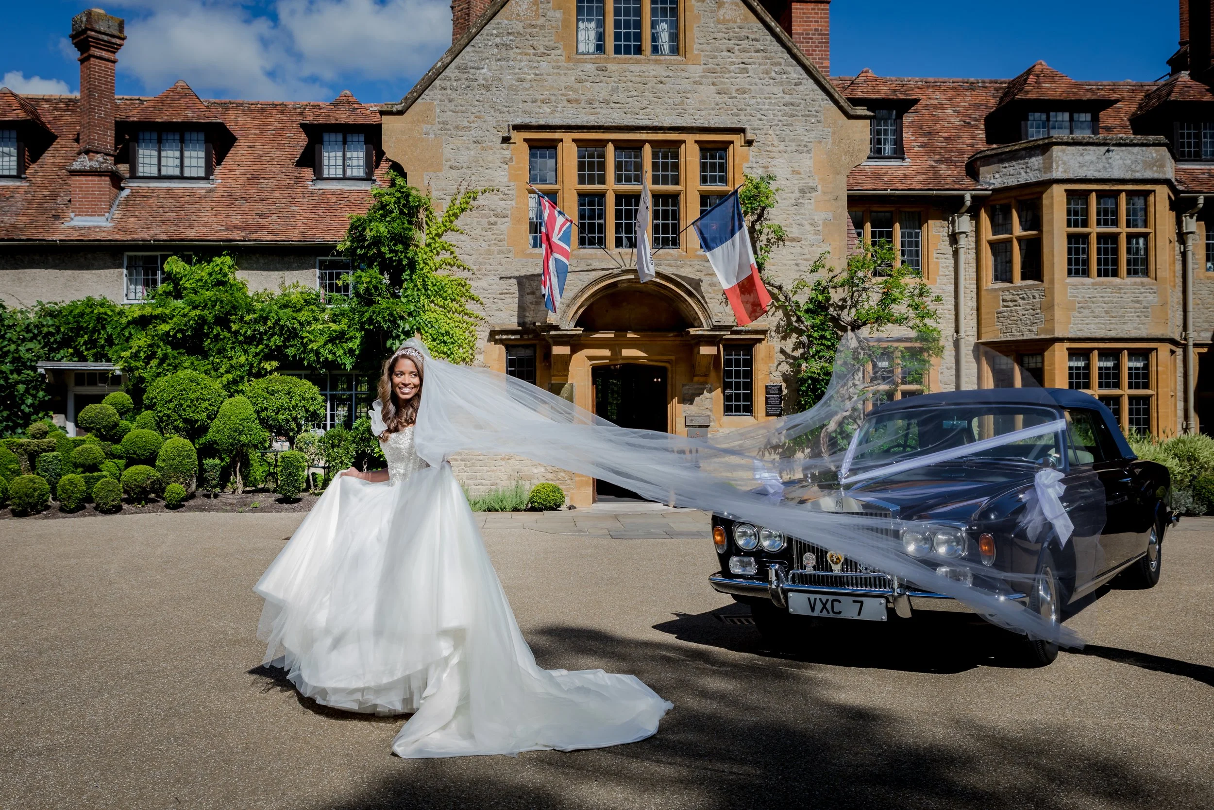 A smiling bride in a wedding gown and tiara, standing outdoors in front of a historic stone building with flags, next to a vintage black convertible car decorated with a white ribbon.