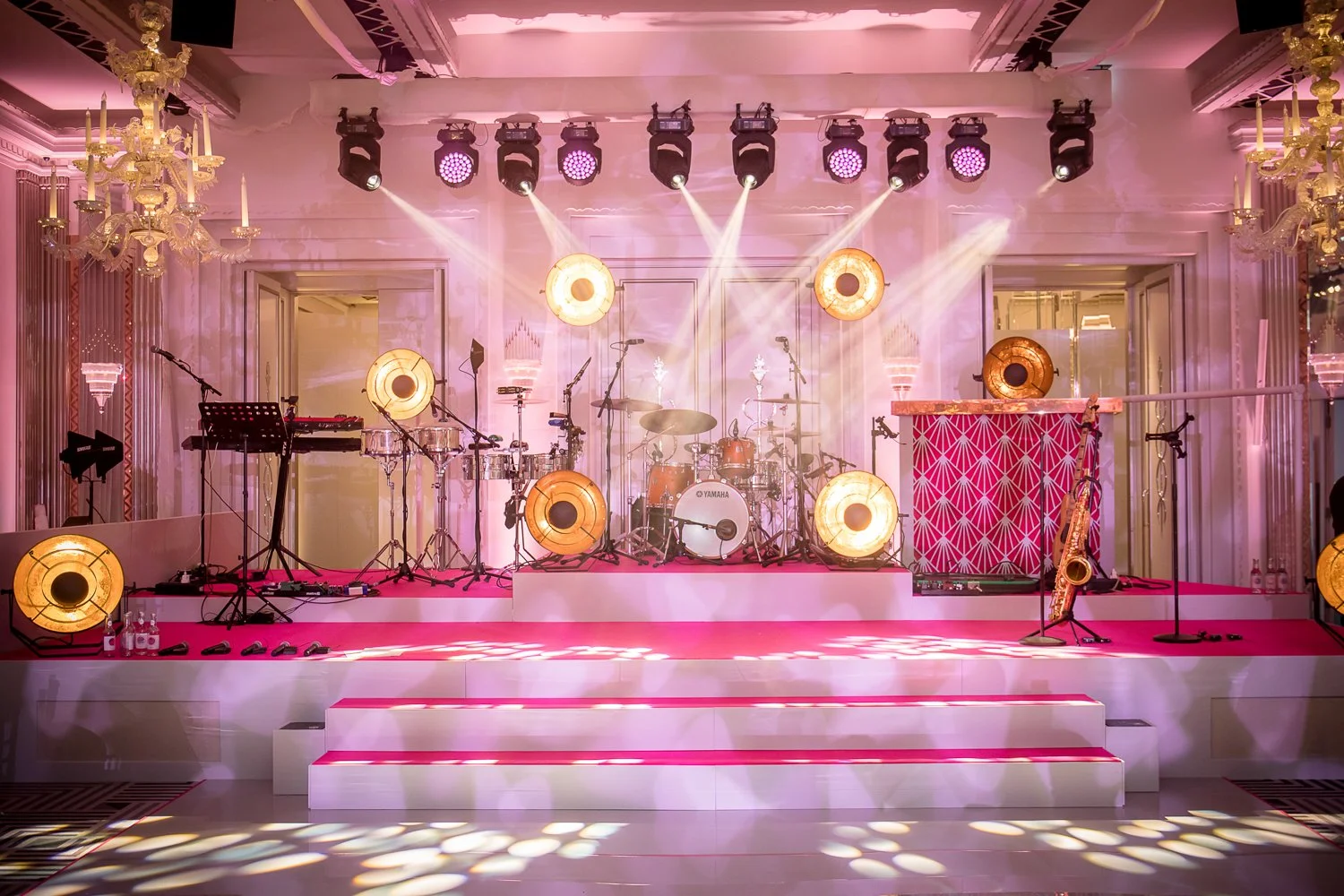 Stage set up for a musical performance with drums, microphones, and instruments, decorated with gold and pink lighting, and chandeliers in an elegant venue.