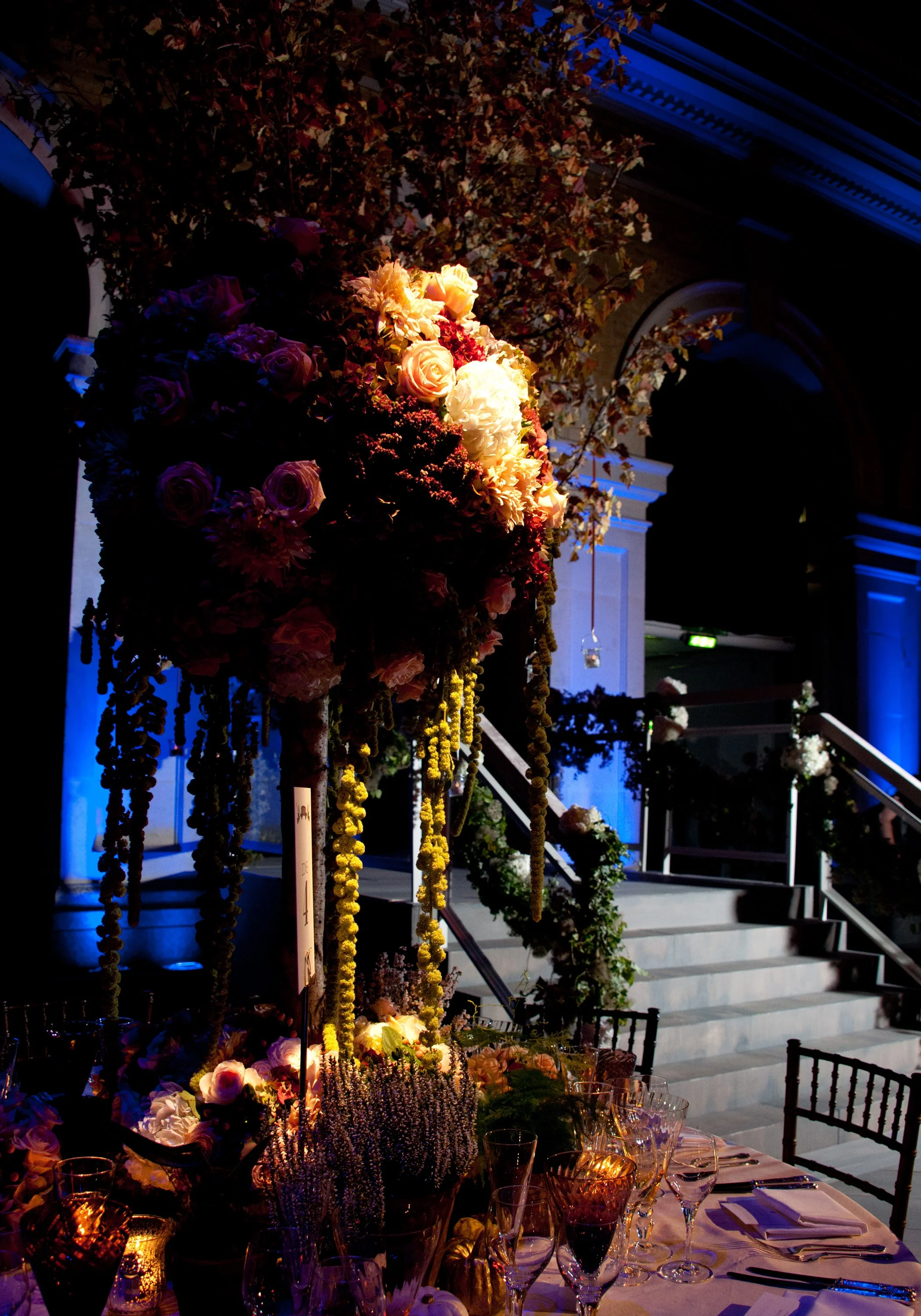 A banquet table decorated with flowers and candles, set for a formal event, with an elaborate floral arrangement in the center. The background shows a staircase and architecture illuminated with blue lighting, indicating an upscale venue.