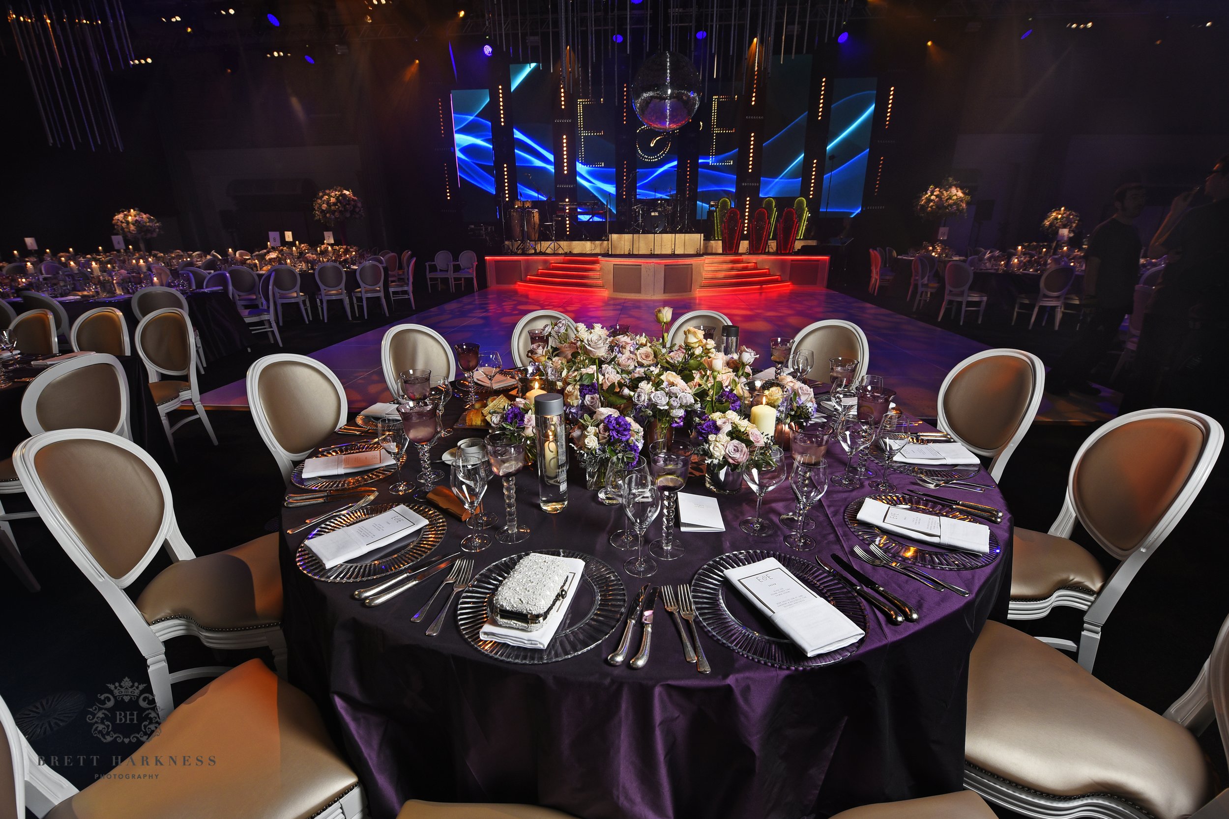 Elegant event venue with round tables covered in purple cloths, set with glassware, silverware, and floral centerpieces, with a stage featuring lighting, large LED screens, and cactus decorations.
