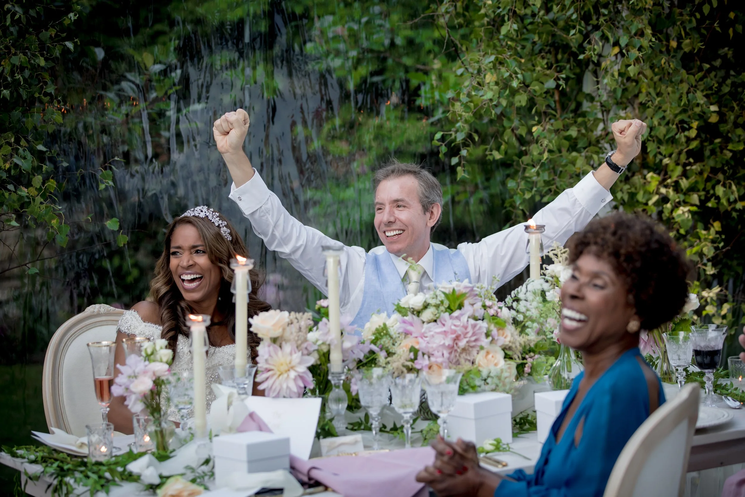 People celebrating at a wedding reception outdoors, with a man in a light blue vest and white shirt raising his arms in celebration, surrounded by women smiling and sitting at a table decorated with flowers and candles.