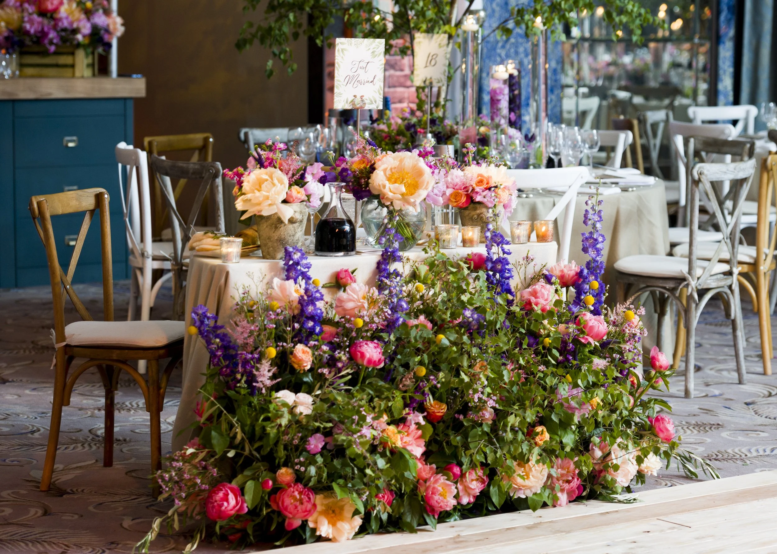 A sitting area decorated with numerous pink, purple, and peach flowers, including tulips and roses, with candles on a beige tablecloth, and signs indicating a wedding celebration.