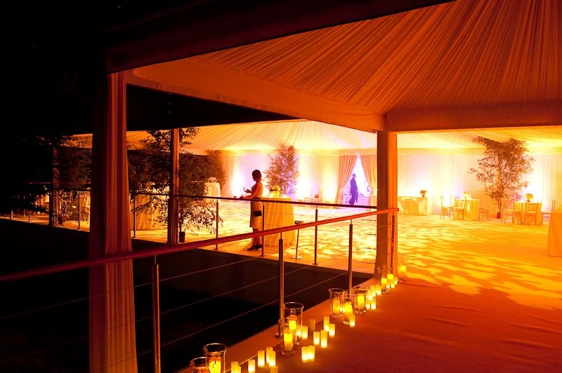 Indoor event space decorated with warm lighting, candles, and potted plants, featuring a woman standing near a railing and another person in the background.