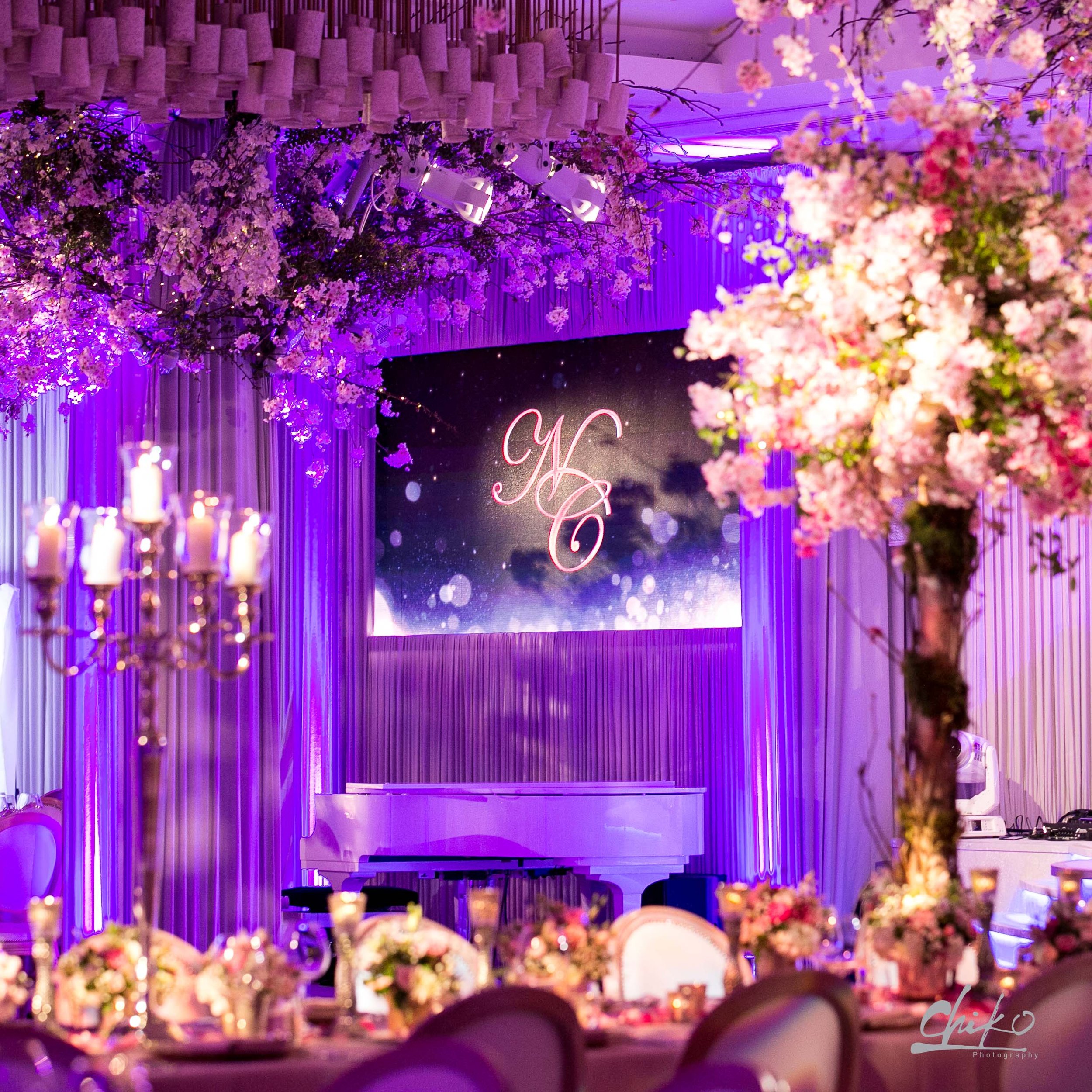 A decorated event space with pink and purple floral arrangements, candles, and elegant table settings, illuminated with purple lighting, likely for a wedding or celebration.