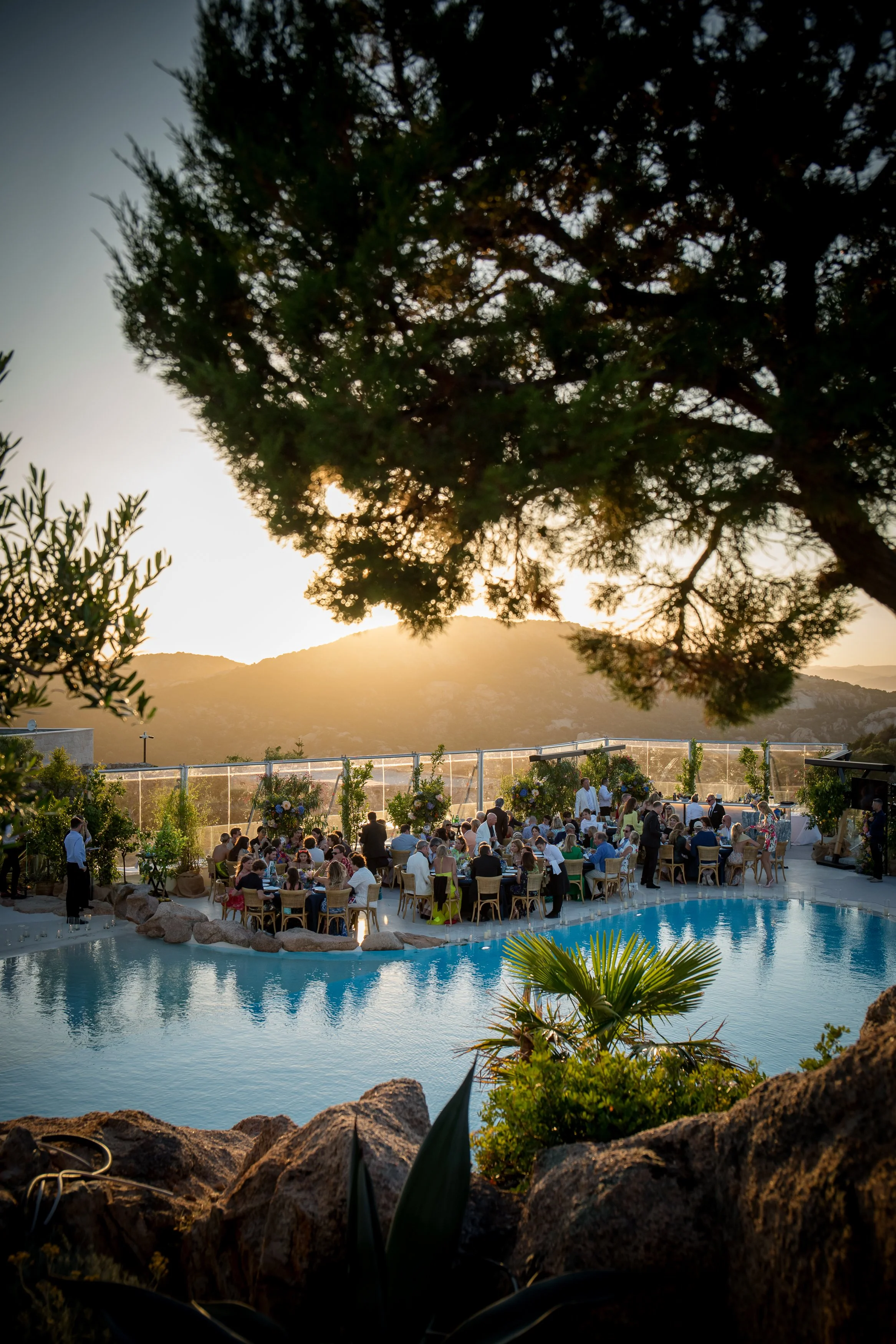At private party in Sardinia where people gathered by a swimming pool during sunset, with trees and mountains in the background.