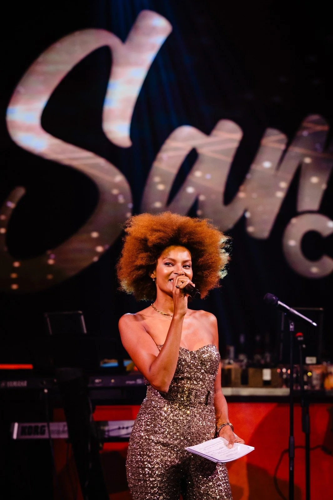 A woman with large curly hair and wearing a sparkly strapless dress holding a microphone and papers, performing on stage with a large illuminated sign in the background.