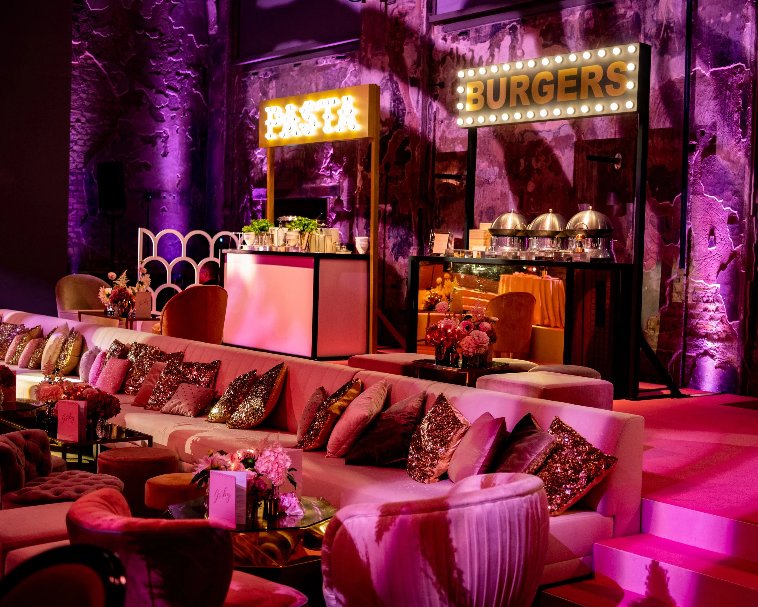 A cozy indoor space decorated with plush pink and gold pillows on white sofas, with flowers on the tables. The background features a textured brick wall with purple lighting, and illuminated signs reading 'PASTA' and 'BURGERS'. BAT MITZVAH AT BATTERS