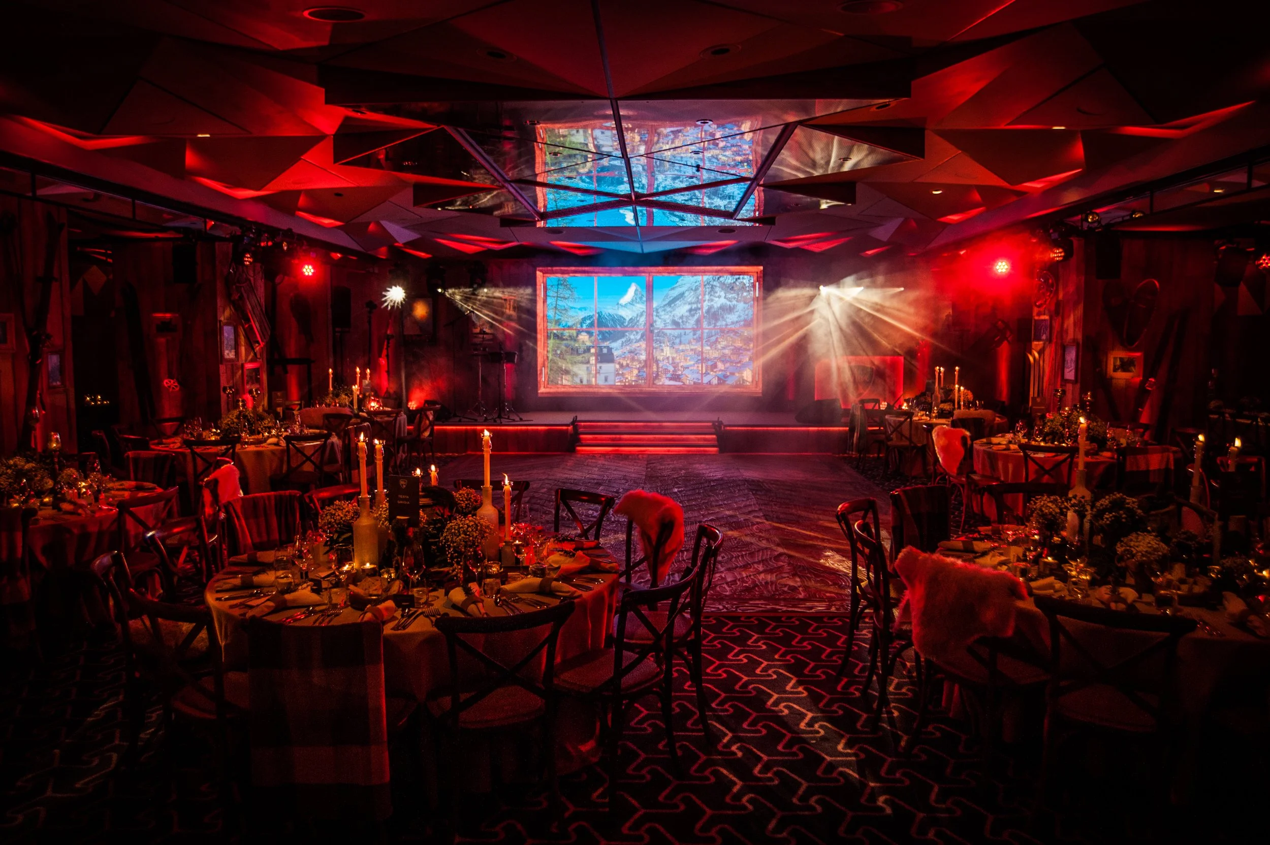 An indoor event space decorated with tables set for a formal dinner, with dim lighting and red and white candles, illuminated by colorful stage lights. A large window scene or backdrop displays a mountain landscape, creating a cozy and elegant ambian