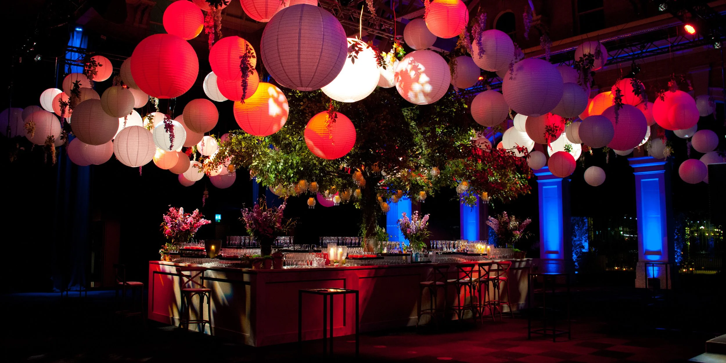 A festive indoor event space decorated with hanging paper lanterns in red, white, and purple hues, with a bar area underneath adorned with floral arrangements and candles. The space features dramatic lighting with blue and purple accents.