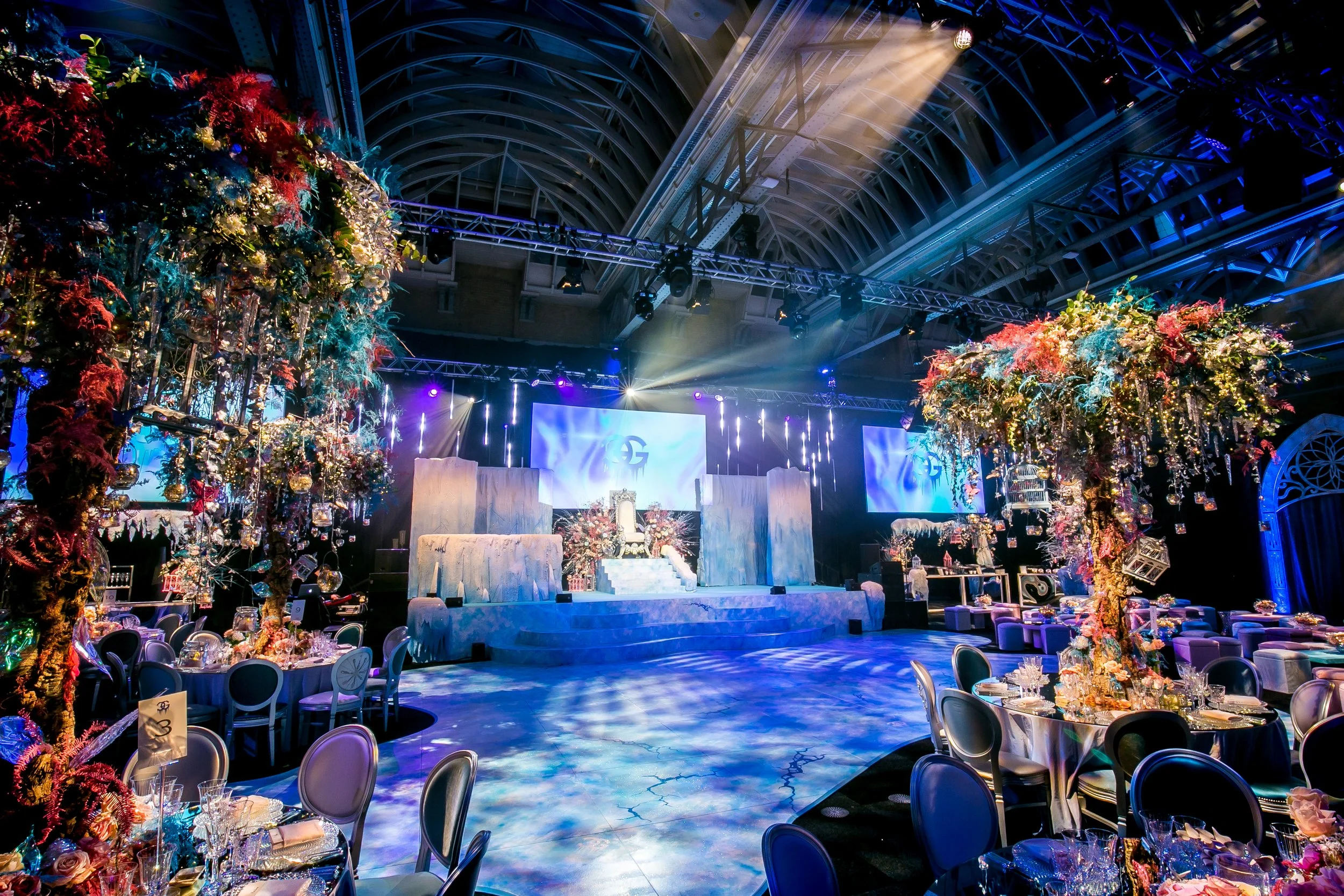 An elaborately decorated event space with floral arrangements, stage lighting, and banquet tables, set up for a celebration or formal event.