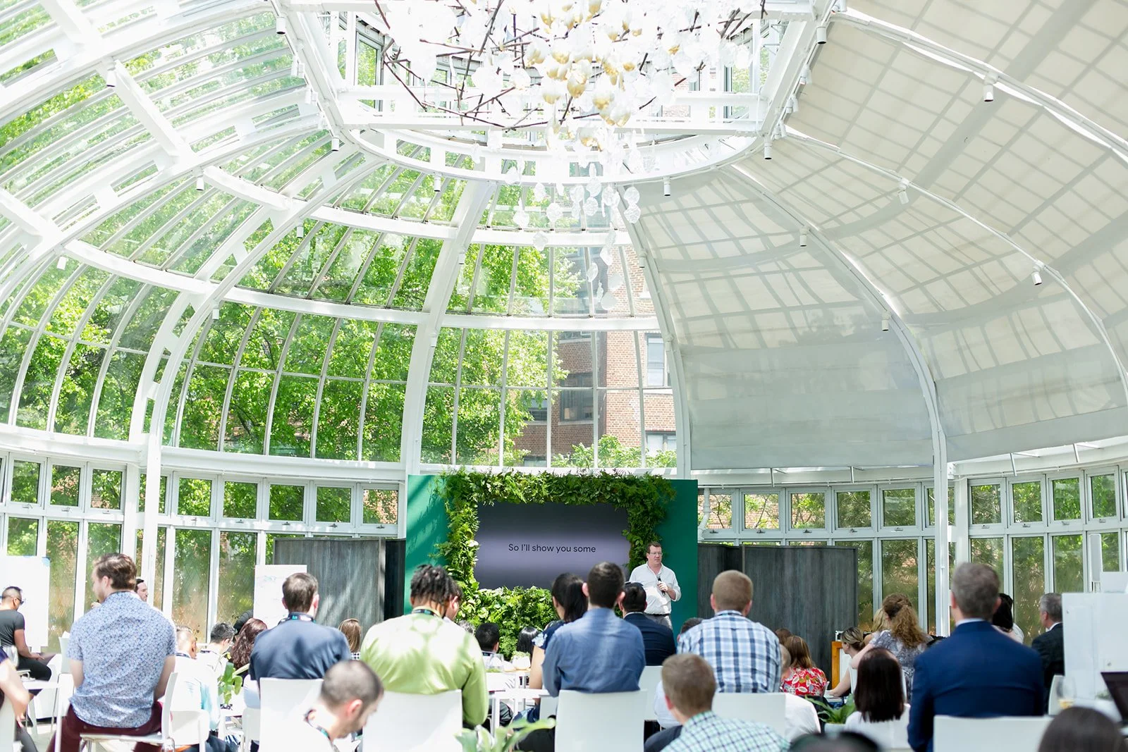 People attending a presentation in a bright indoor greenhouse-like space with large glass windows and a high arched ceiling, while a speaker stands at the front near a screen.