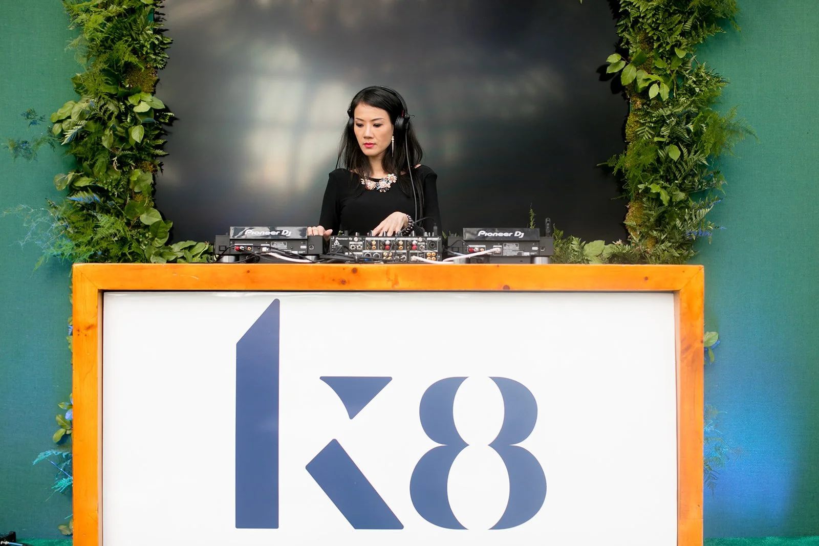 A female DJ wearing a black dress and headphones stands behind a DJ booth with Pioneer DJ equipment, set against a backdrop of green foliage and a large black screen, at an event marked with the number 128.