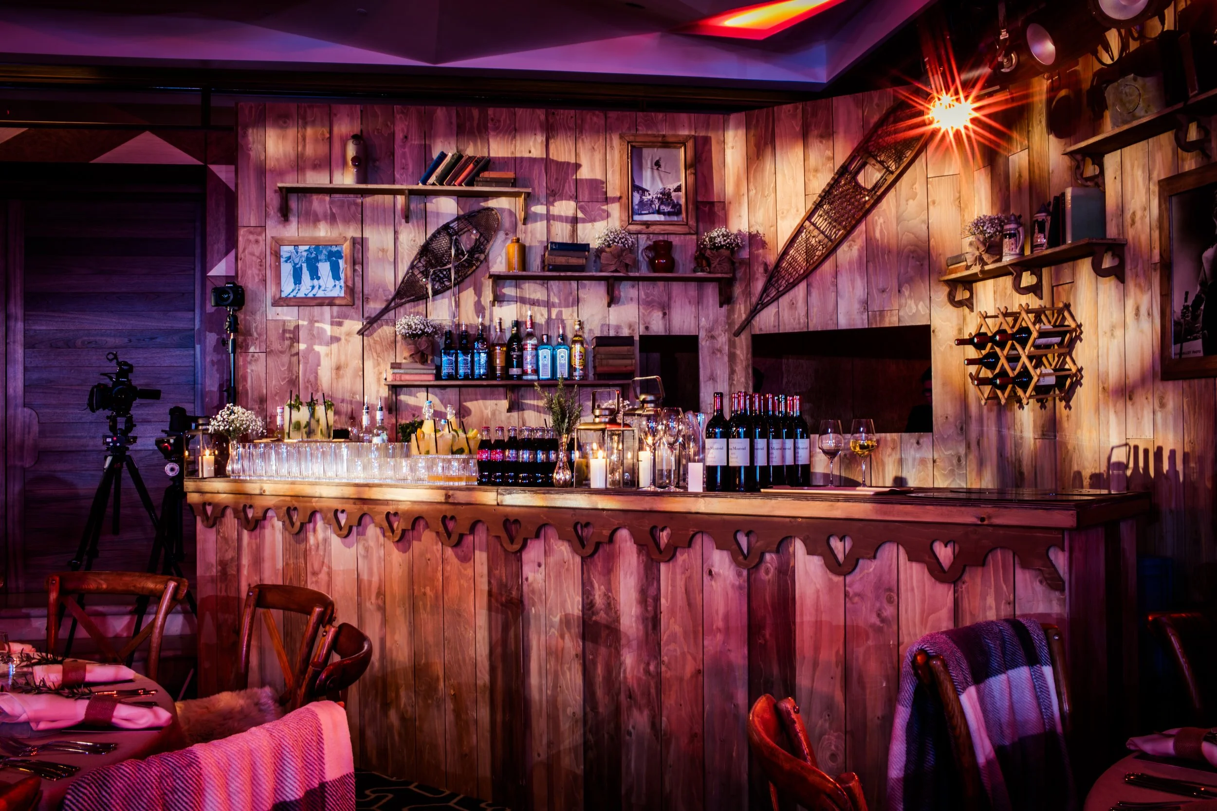 A cozy dinner setup in a rustic wine bar with wood-paneled walls, shelves with books and decor, a bar area with various alcohol bottles, glasses, candles, and wine bottles. There are vintage skis hanging on the wall, a camera on a stand, and warm, am