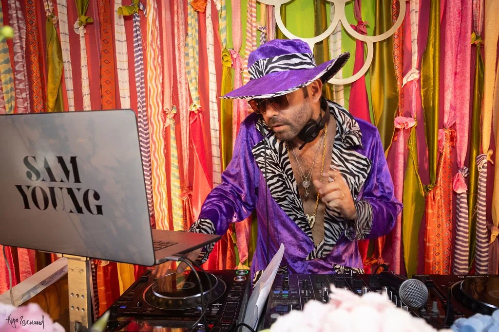 Man in a purple velvet outfit and wide-brimmed purple hat with zebra pattern, wearing sunglasses, headphones, and jewelry, DJing at a colorful event with a multicolored fabric backdrop.