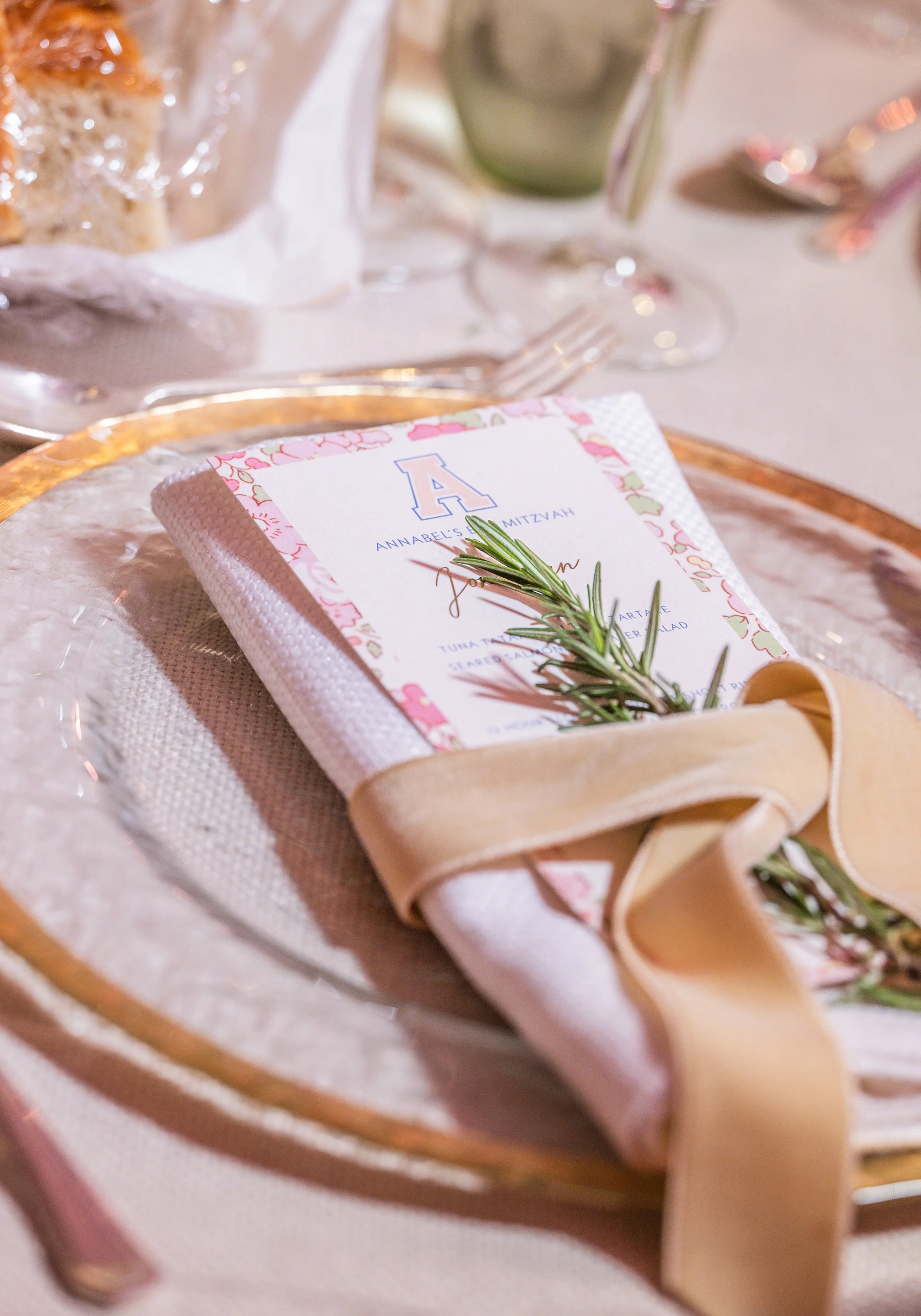 A close-up of a table setting for a special occasion, featuring a decorative napkin with the letter 'A' and the words 'Annabel's Mitzvah,' garnished with a sprig of rosemary, surrounded by glassware, a cake, and utensils with a blurred background.