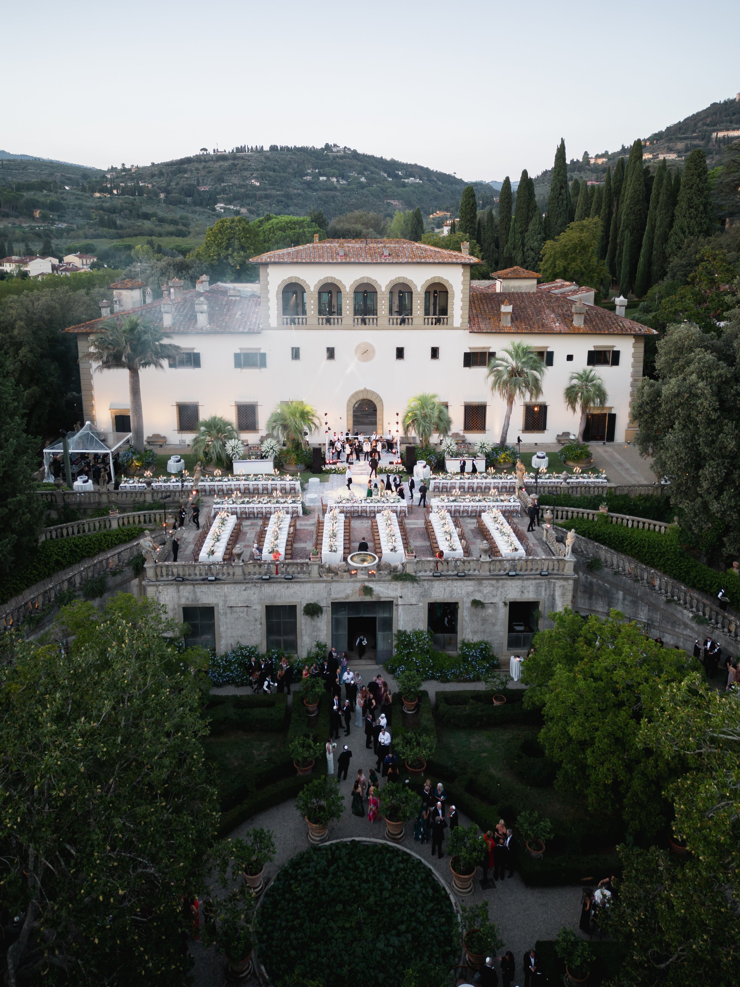 Wedding reception at Villa Palmieri in Tuscany. Grand white building with tanned roof tiles and greenery. Long tables shown from an ariel view. 