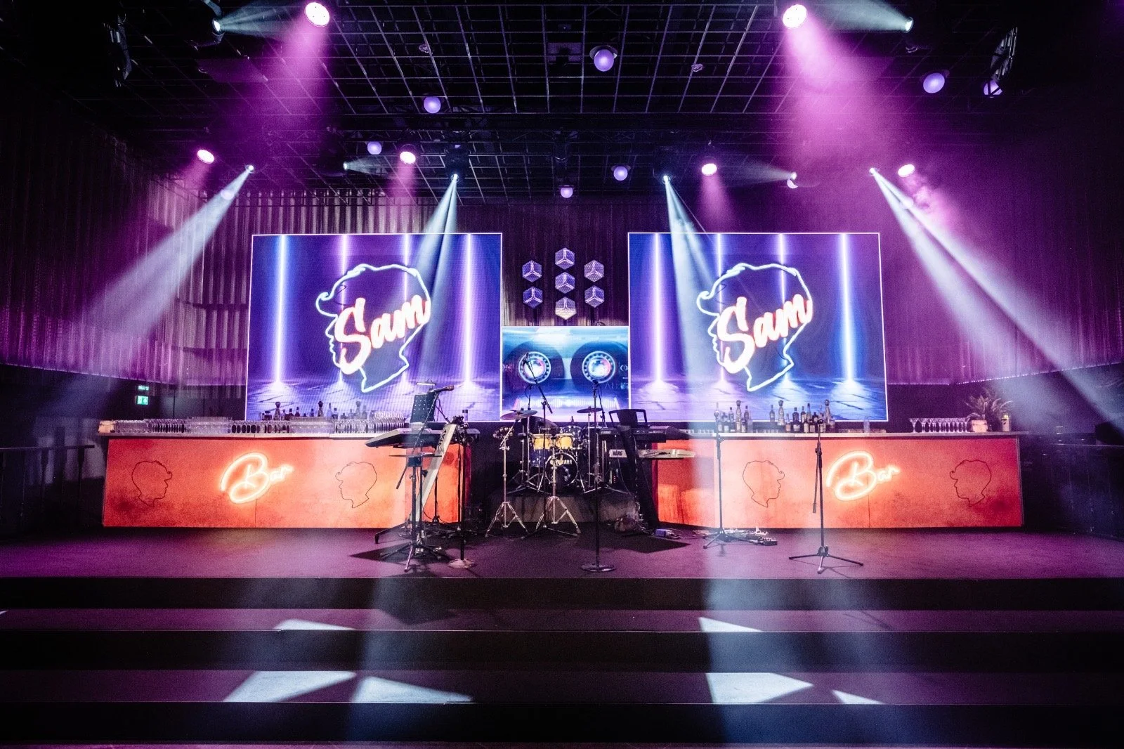 Empty stage with musical instruments under purple lighting, large screens displaying a woman's silhouette with the name 'Sam', bar area with neon sign 'Bar' and cocktail glasses, and bar setup with bottles and glasses.