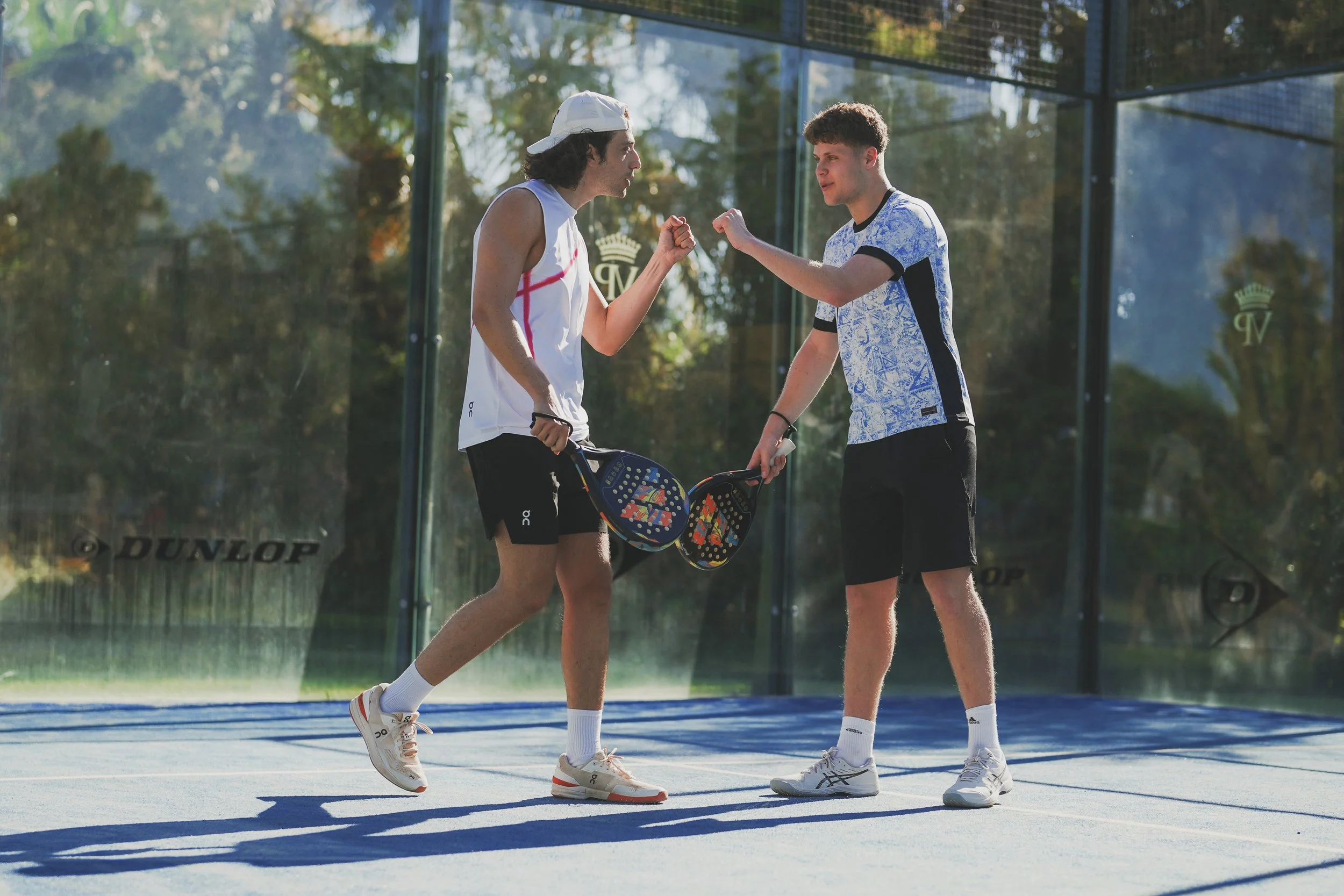 Two young men playing pickleball on an outdoor court, fist bumping each other, with paddle rackets in hand, wearing athletic clothing and sneakers, surrounded by trees and fencing.