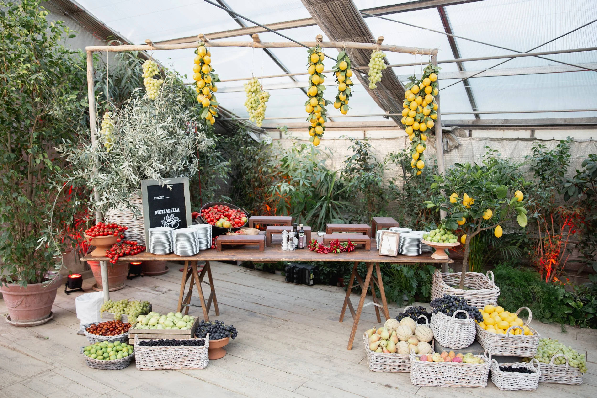 A fruit display table with various baskets of grapes, apples, and other fruits, under a greenhouse with hanging lemons and a small chalkboard sign that says 'Mozzarella'. Venue is in Tuscany at Serre Torrigiani Bistrot