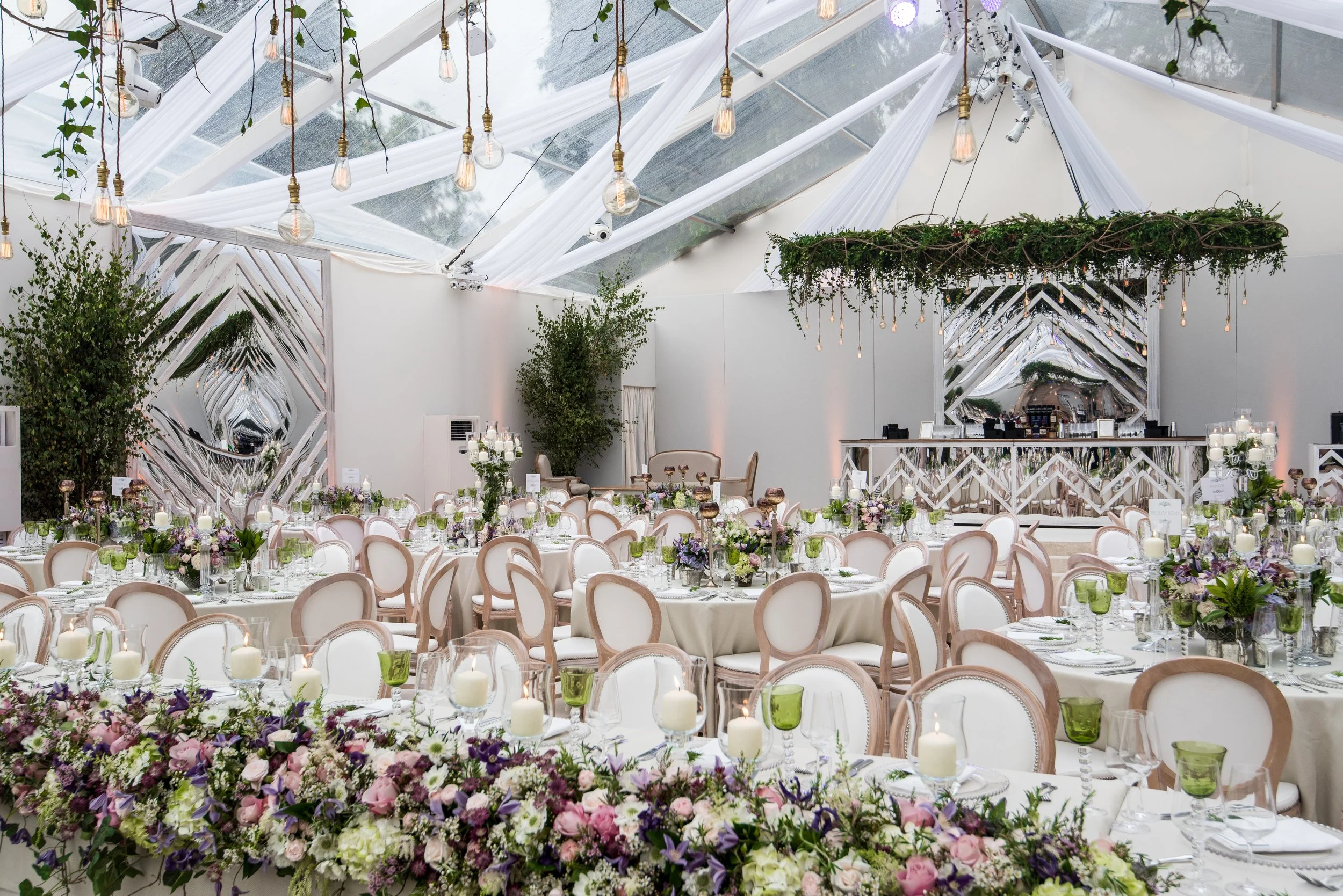 Elegant indoor event space decorated with white drapery and hanging Edison bulb lights, featuring tables set with floral centerpieces, candles, and glassware, and large ornamental mirrors on the walls.