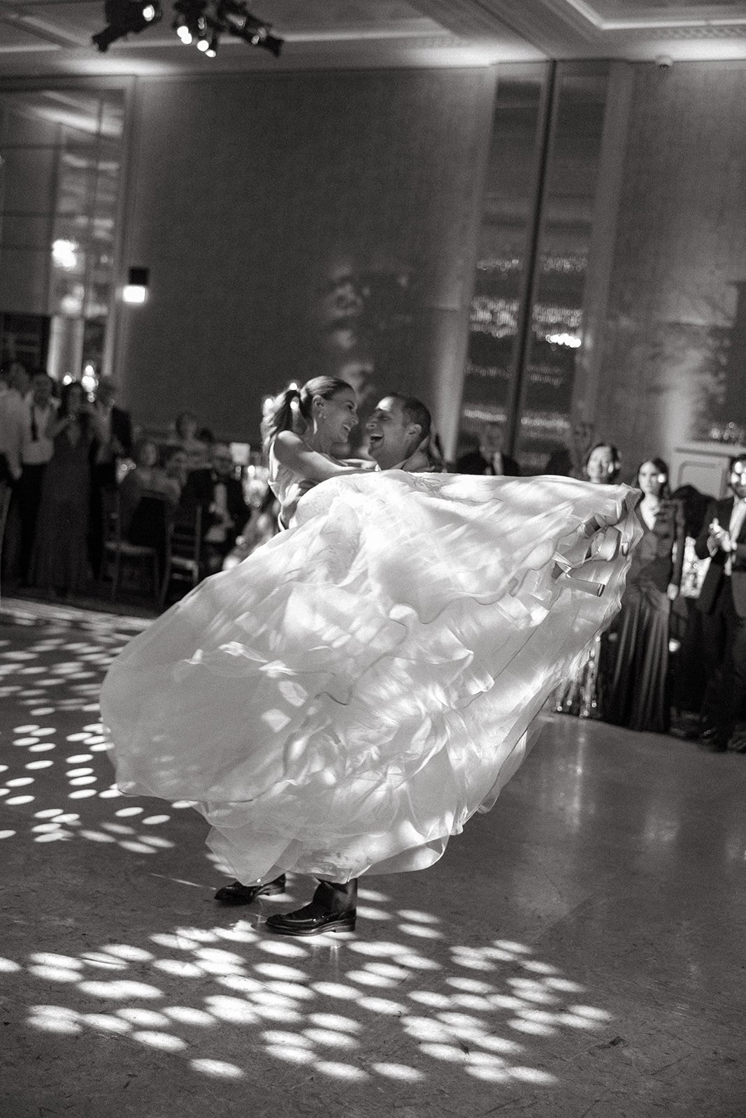 A bride and groom dancing at their wedding reception, with the groom lifting the bride. The bride's dress is flowing, and they are smiling. The reception area is dimly lit with spotlights and guests watching in the background at The Peninsula London