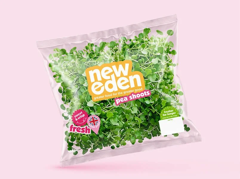 Green pea shoots in a clear plastic bag with labels indicating they are fresh and new eden brand, on a pink background.