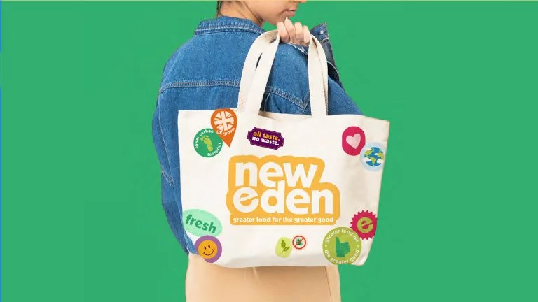 Person holding a reusable shopping bag with this logo for New Eden, featuring colorful stickers and tags on a green background.
