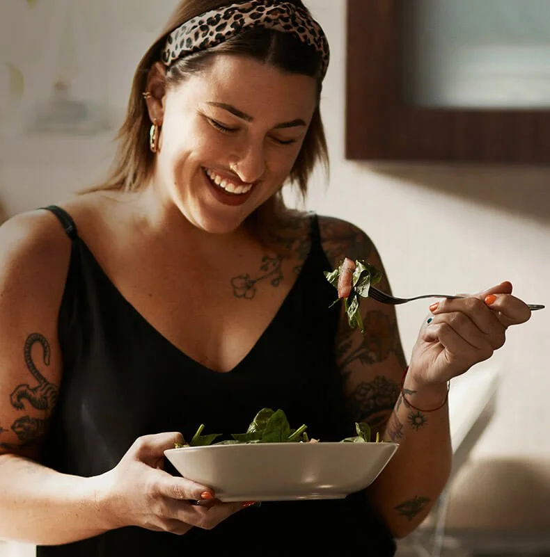 A woman with tattoos and a leopard print headband is smiling and enjoying a salad, holding a white bowl in one hand and a fork with salad in the other.