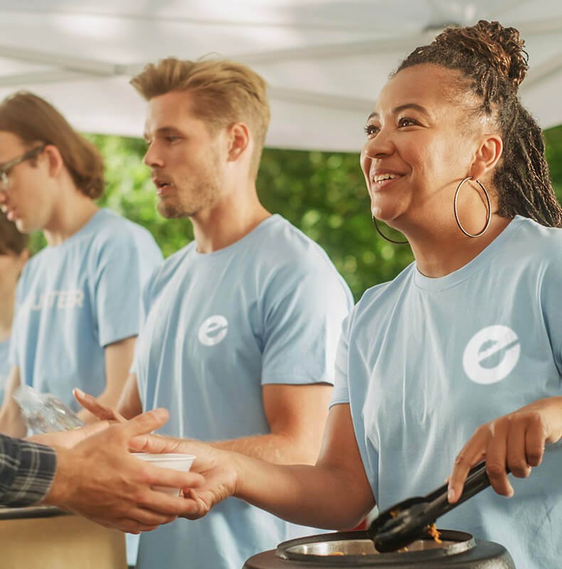 People in light blue shirts participating in a community service event, with a woman handing a meal to someone outside the frame.