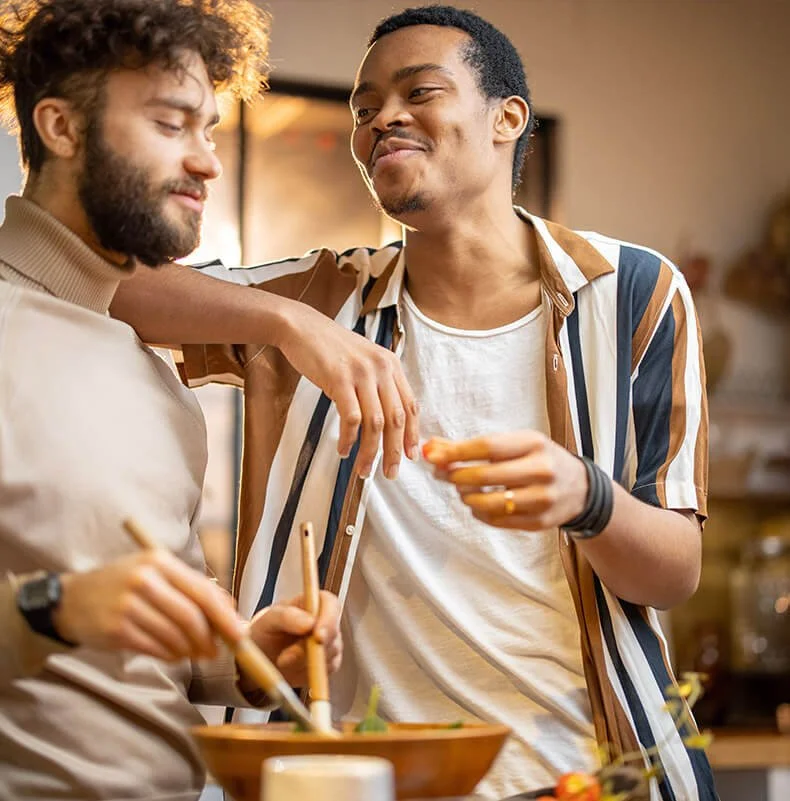 Two men enjoying a meal and conversation at a restaurant or cafe, one with curly hair and beard, the other with short hair, wearing a striped shirt.