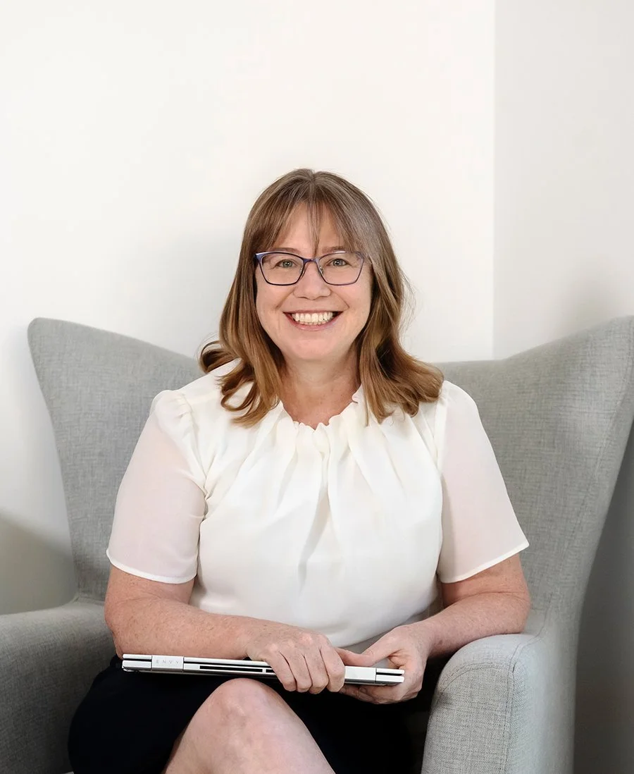A woman with glasses and shoulder-length hair sitting in a light gray armchair, smiling and holding a closed silver laptop.