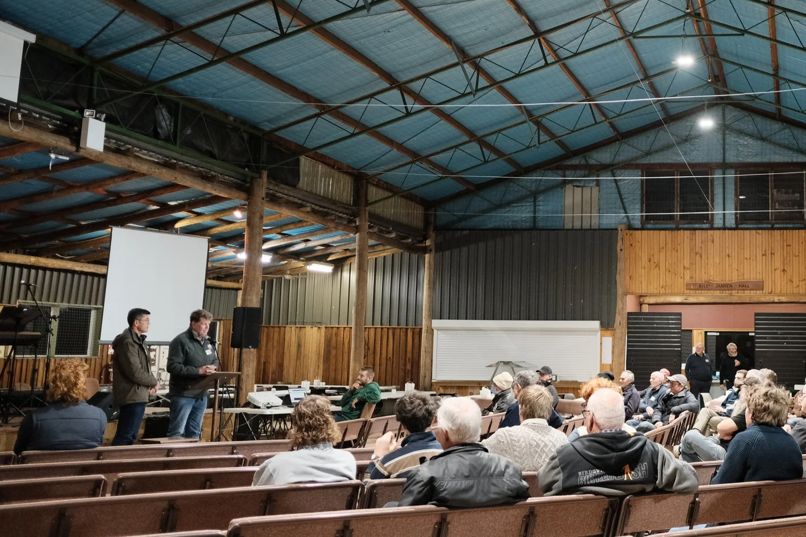 People attending a meeting or presentation inside a barn or community hall with wooden walls and a high, blue metal roof. Two men are standing at a lectern at the front, speaking to the audience, with a white projection screen behind them. Audience members are seated, some taking notes. There is sound equipment and a piano on the left side.