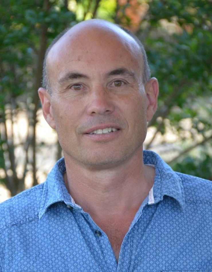 A middle-aged man with a bald head and light skin, wearing a blue button-up shirt, standing outdoors with trees and greenery in the background.