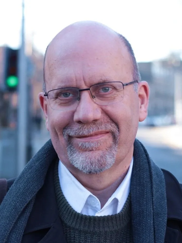 A close-up photo of a smiling middle-aged man with glasses and a goatee, wearing a white shirt, dark sweater, and a dark coat, outdoors with a blurred urban background and a green traffic light visible.