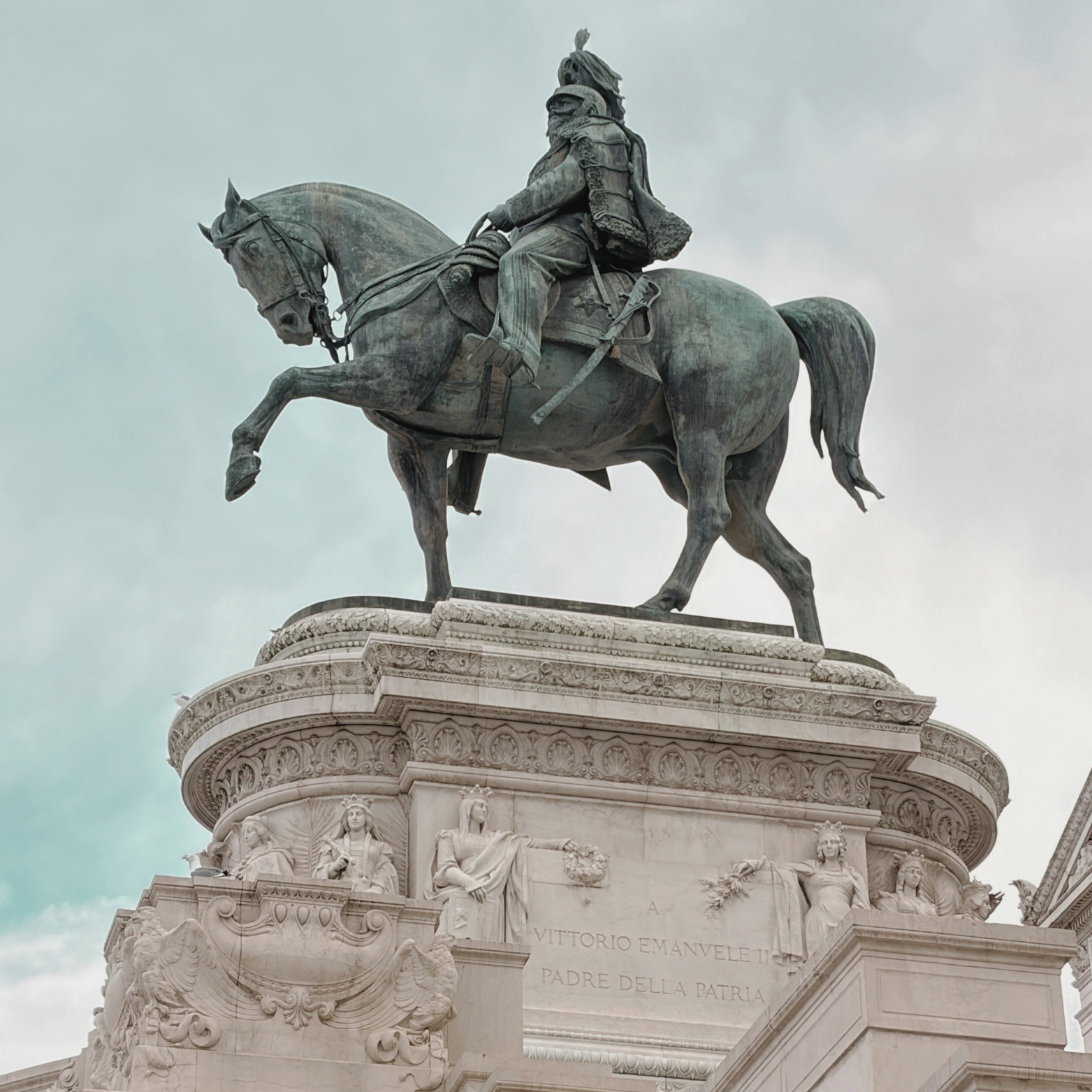 A large bronze statue of a man on a horse, placed on top of a decorative stone pedestal with carvings and Latin inscriptions, against a cloudy sky.