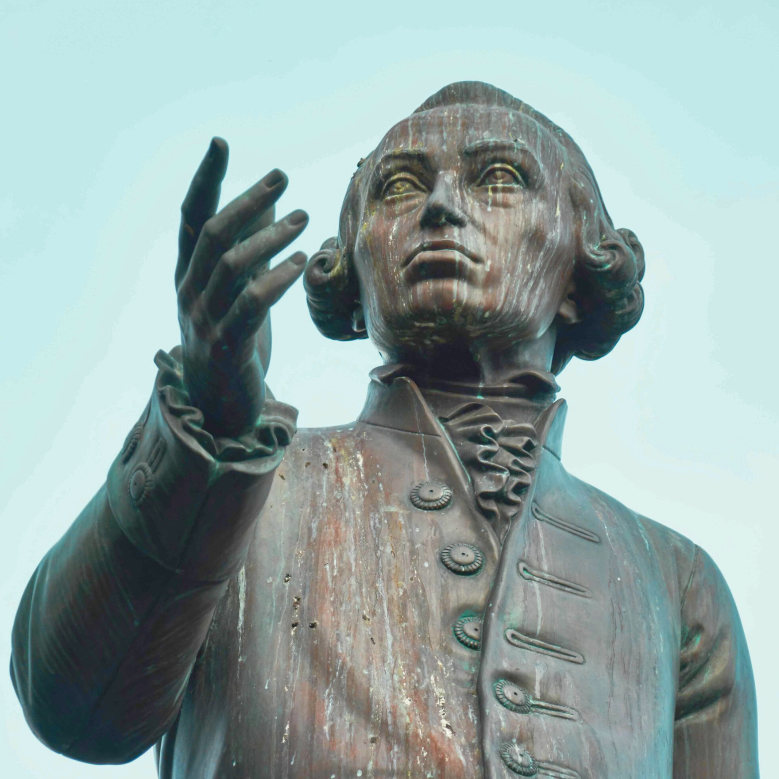 Close-up of a bronze statue of a historical figure with detailed facial features, wearing a colonial-era coat with large buttons, and gesturing with his right hand against a pale sky background.
