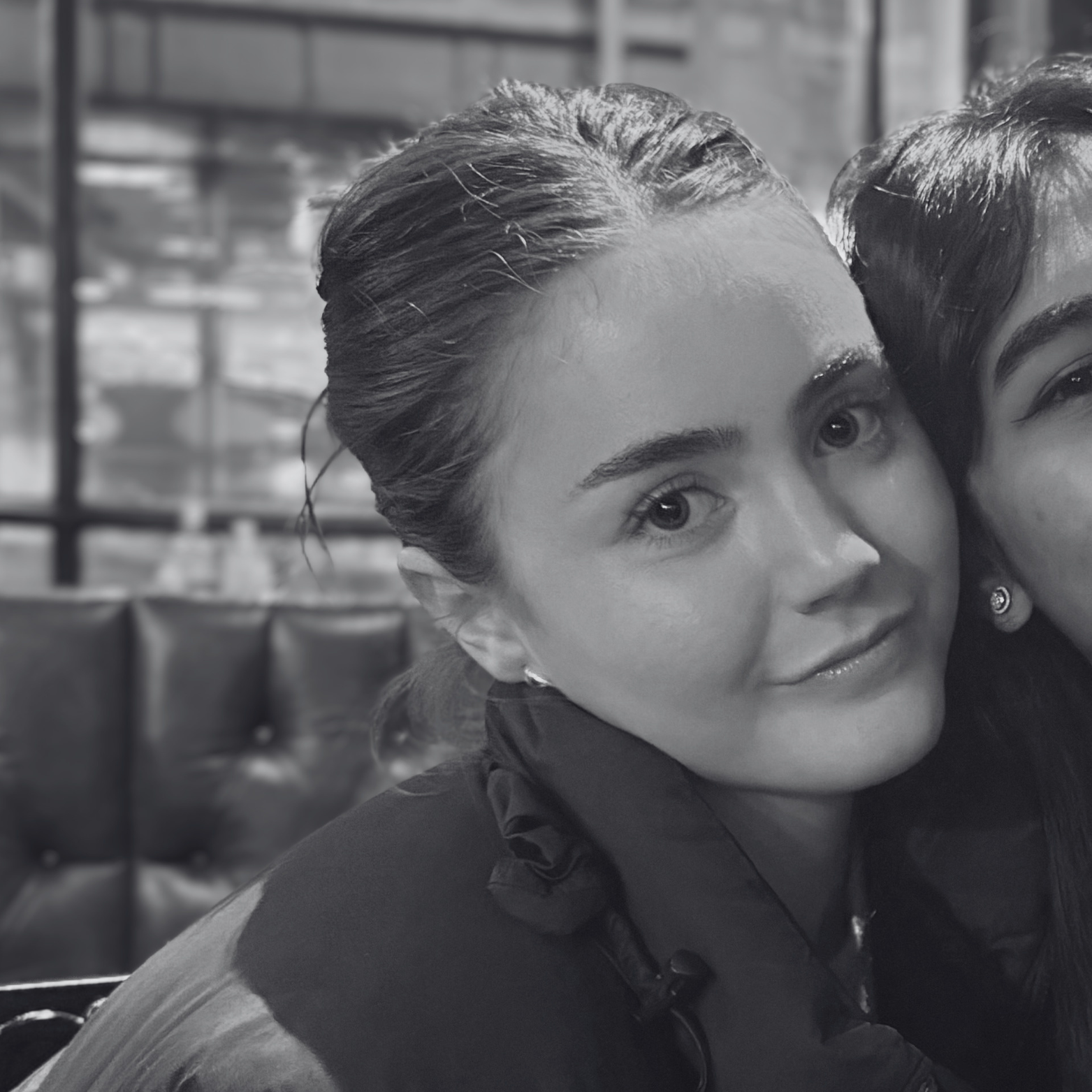 Close-up of a woman with dark hair smiling in black and white, with a person partially visible beside her. Background appears to be a cafe or restaurant setting.