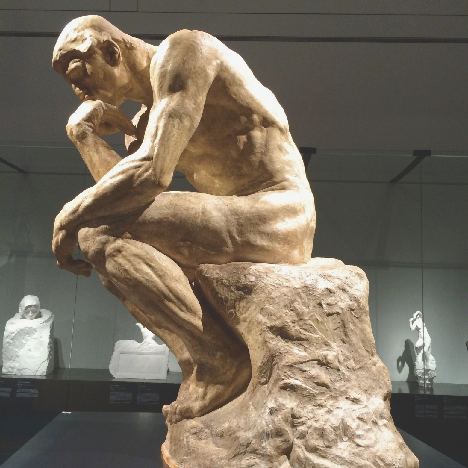 A stone sculpture of a man sitting on a rock with his chin resting on his hand, in a contemplative pose, displayed in a museum.