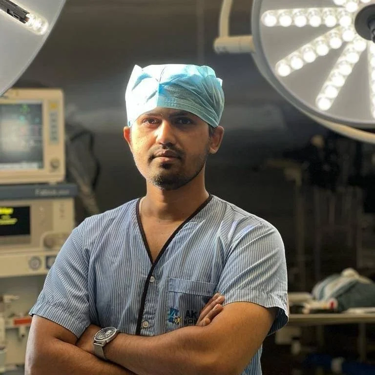 A surgeon in scrubs and a surgical cap posing in an operating room with medical equipment and bright surgical lights.