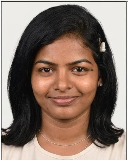 Portrait of a woman with black hair, a pearl hair clip, a small facial piercing, wearing a light-colored top and a delicate necklace, against a plain background.