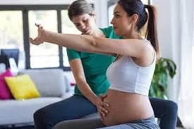 Pregnant woman doing a shoulder stretch with assistance from a woman in a bright, modern living room
