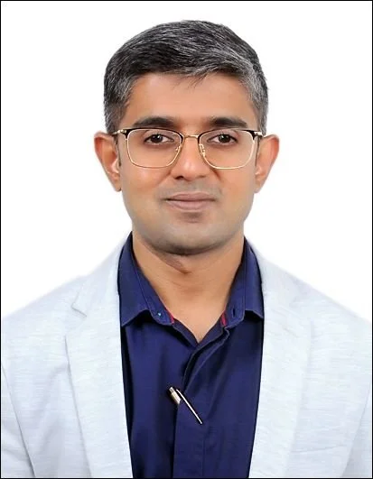 Portrait of a man with short gray hair, glasses, wearing a light gray blazer and navy blue shirt against a white background.