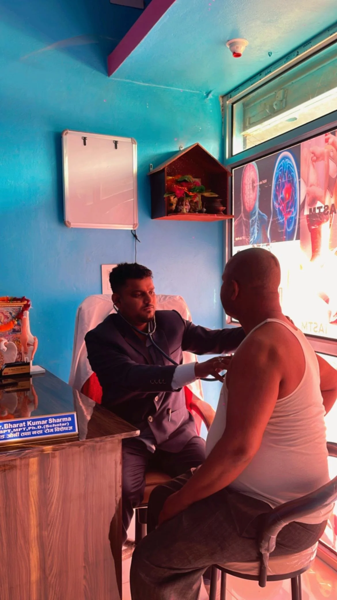 A doctor examining a man in a clinic with a colorful interior, a blue wall, a whiteboard, and a window with a digital display.